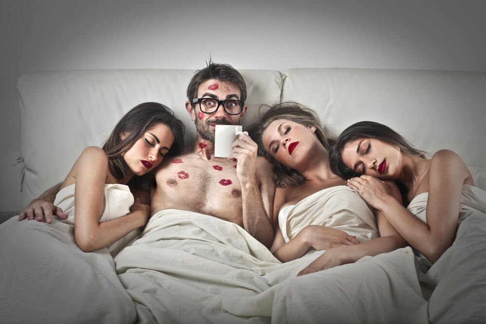 Ein Typ mit drei schönen Frauen, die im Bett liegen. Der Mann hat Kussmarken auf Gesicht und Brust.